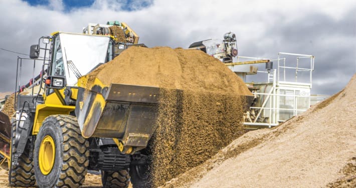 Dymax sand and gravel bucket on Komatsu loader. Sand is spilling over front edge of bucket. Sand pit in background.